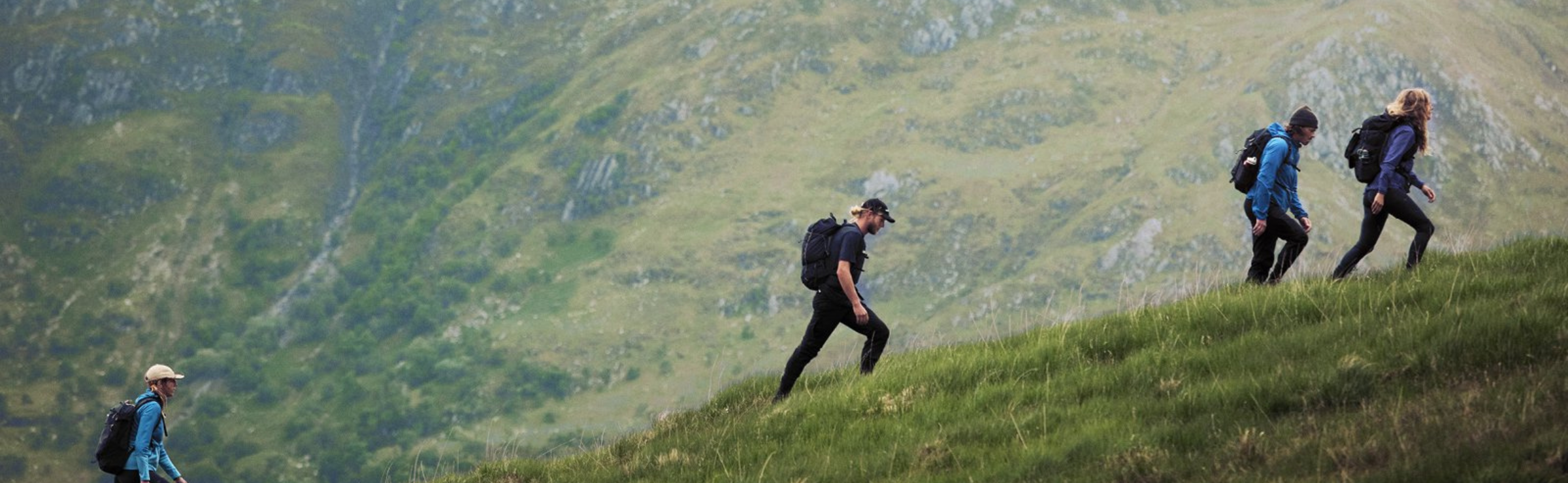 Four hikers with backpacks walking on a grassy hillside with a mountainous background.